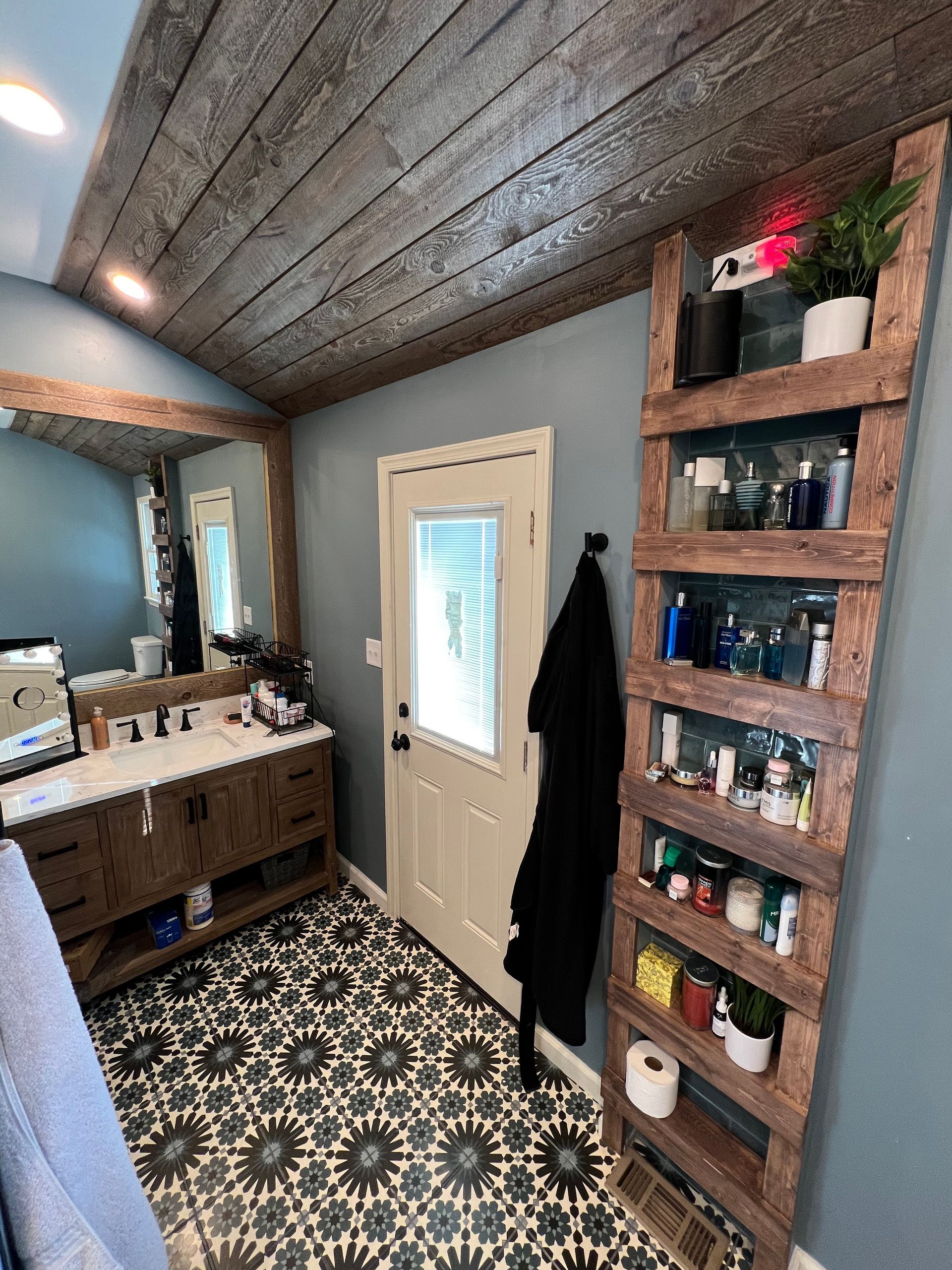 Bathroom with wood shelving, patterned floor, vanity, and door. Blue walls, wood ceiling.