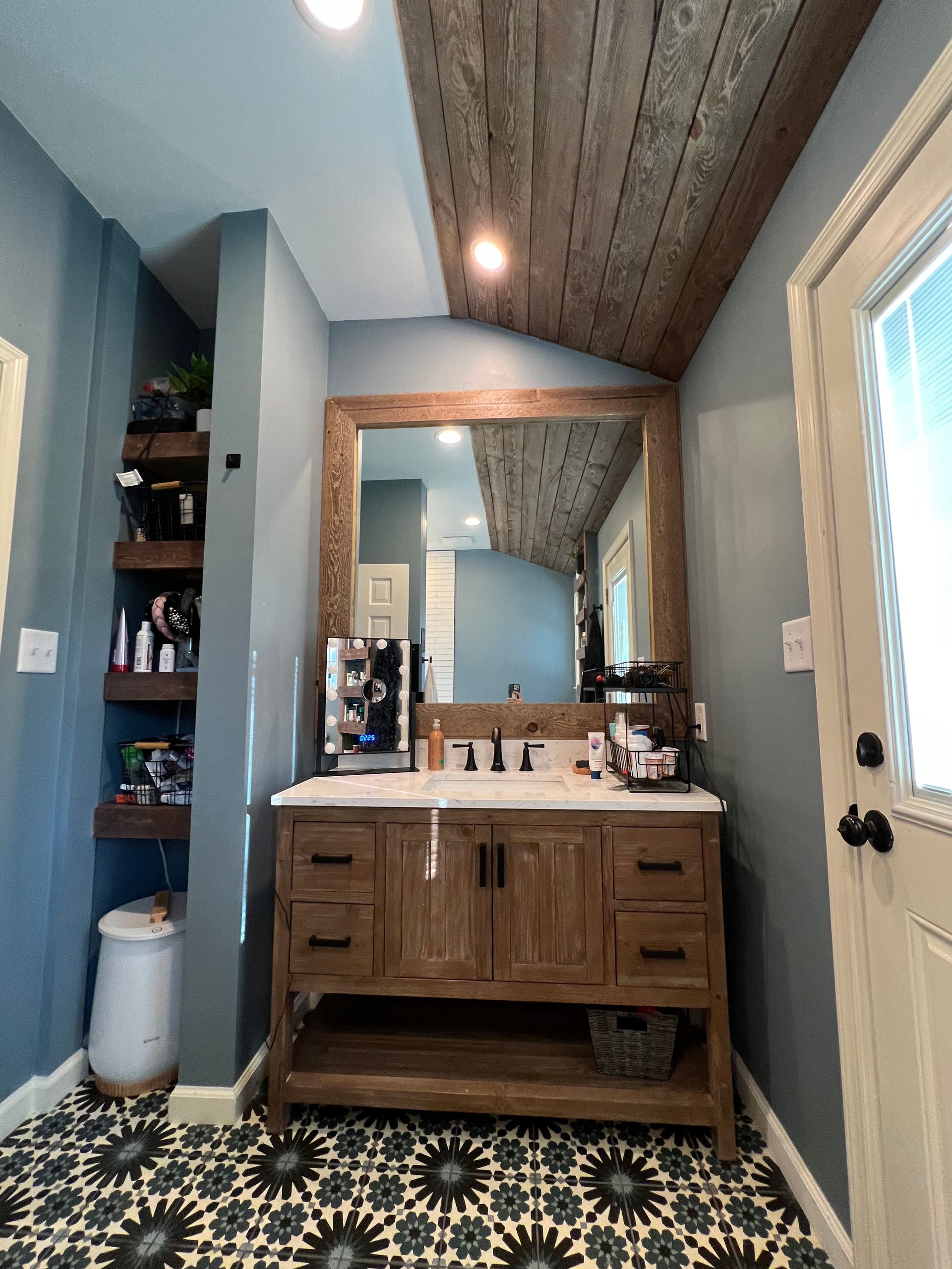 Bathroom with wooden ceiling, vanity, patterned floor, and open shelving on a blue wall.