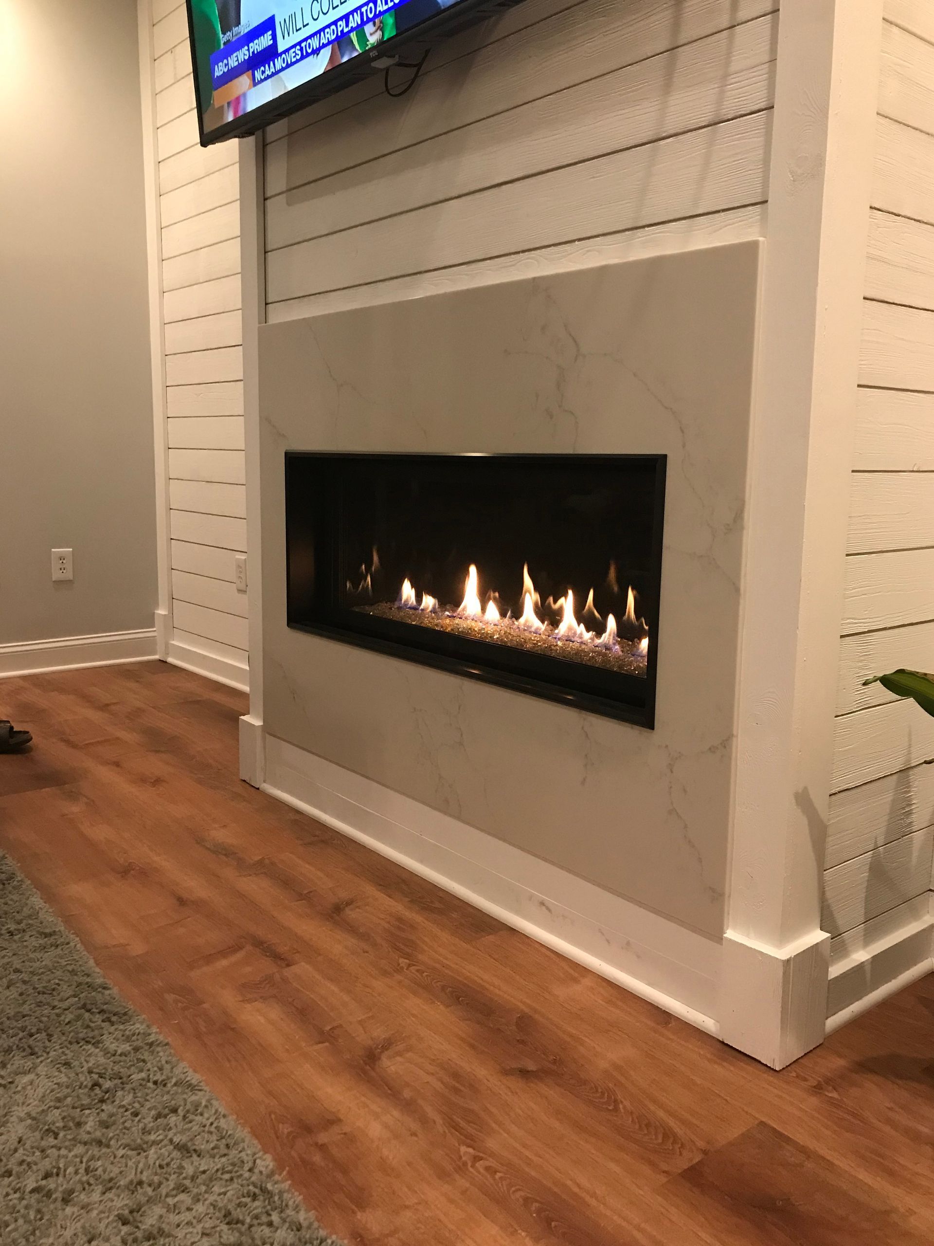 A modern fireplace with a glowing fire, mounted on a white shiplap wall with a TV above it.