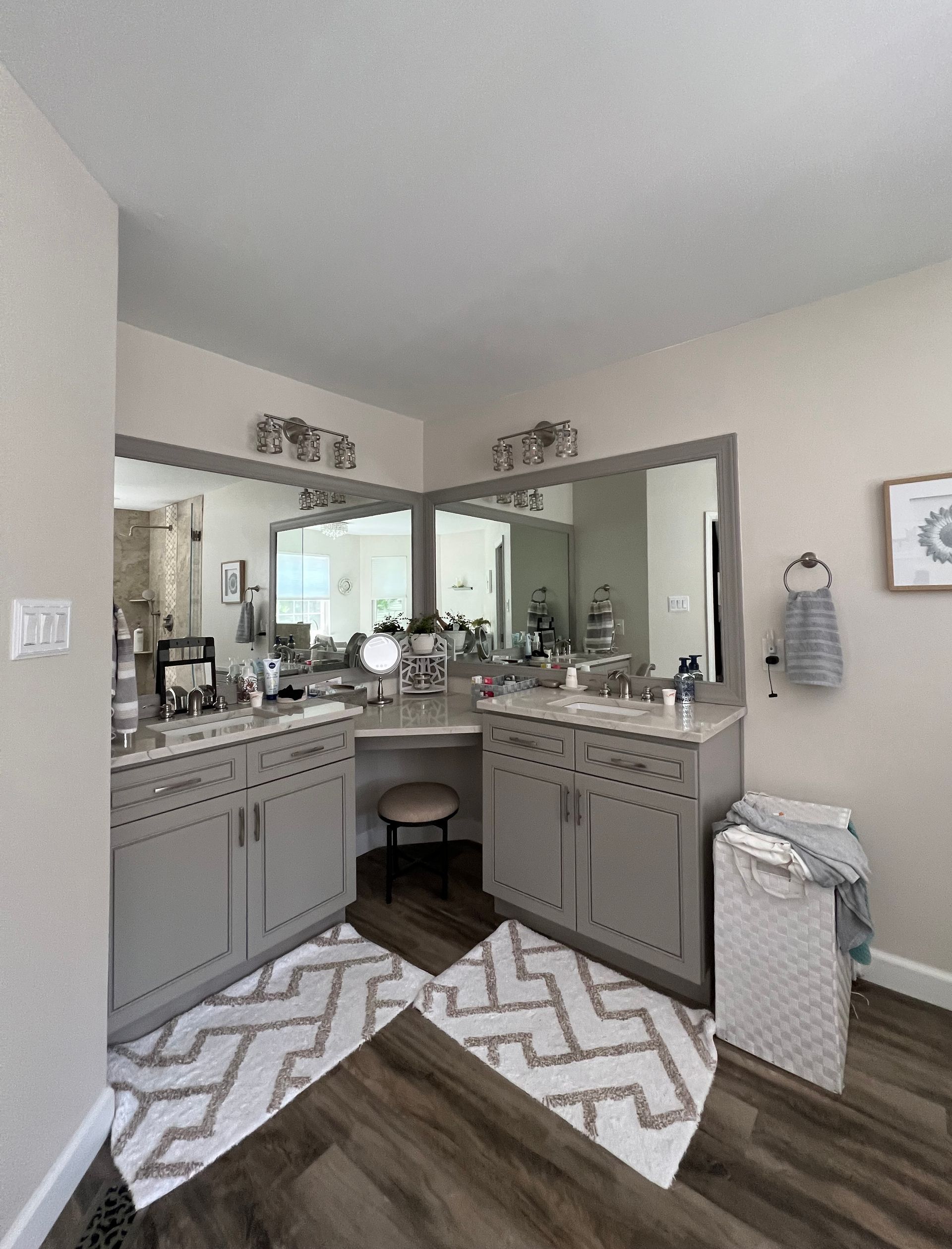 A modern bathroom with gray cabinets, corner vanity, large mirrors, and patterned rugs.
