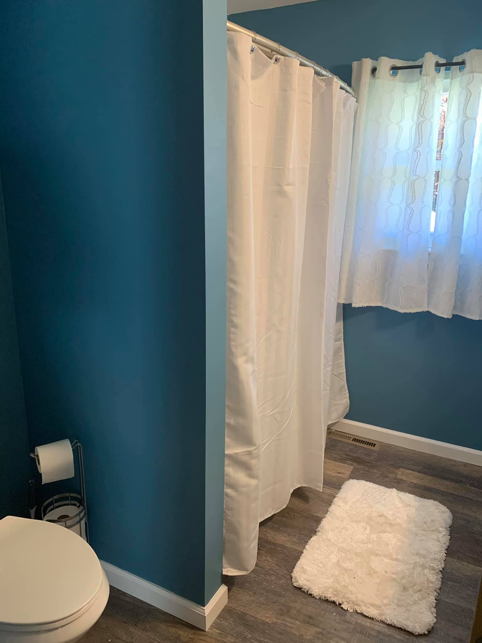 A bathroom with a toilet , shower curtain , rug and window.