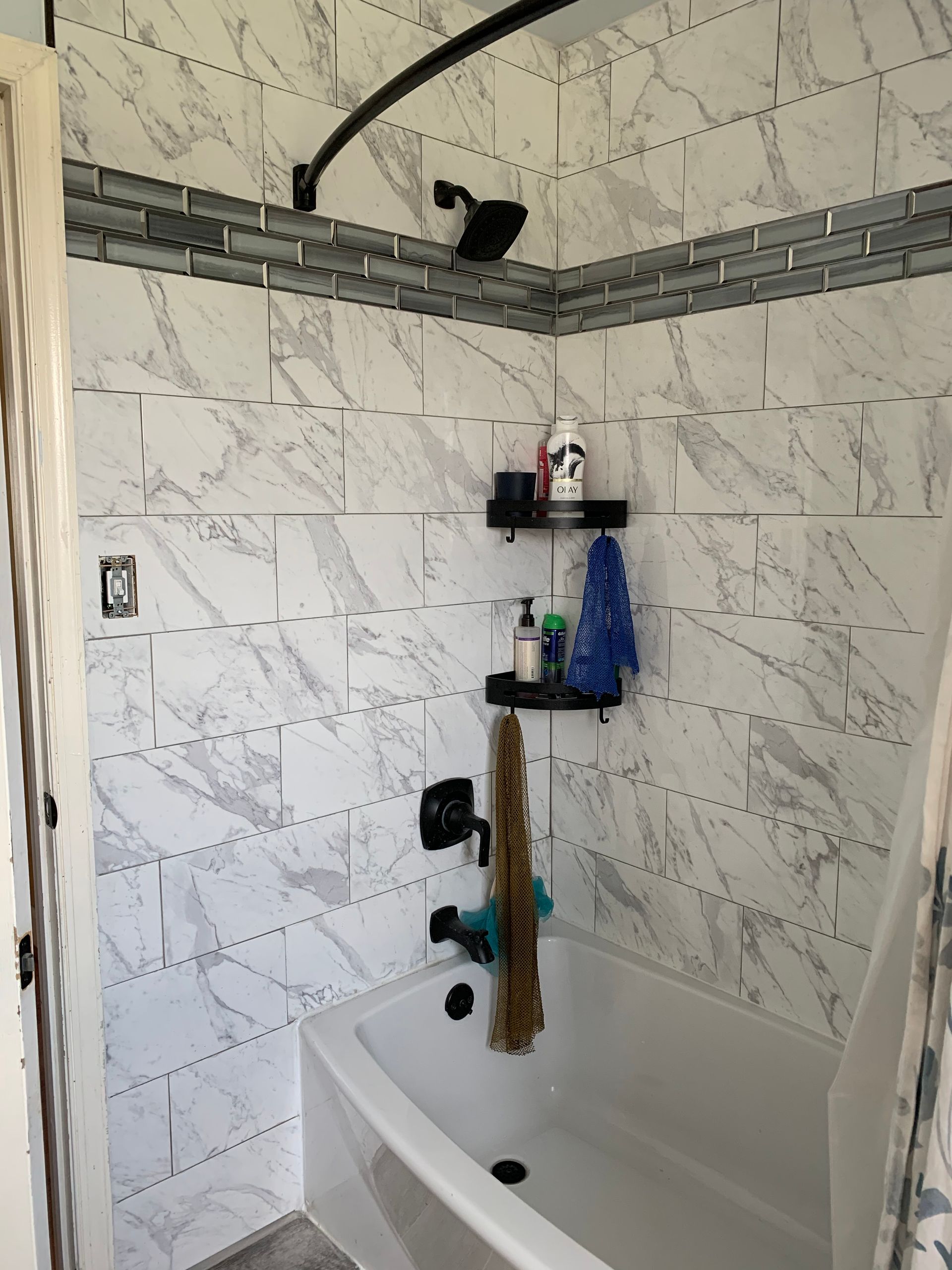 Bathroom with white marble-look tile, gray accent band, black showerhead and curved rod, toiletries on a shelf.