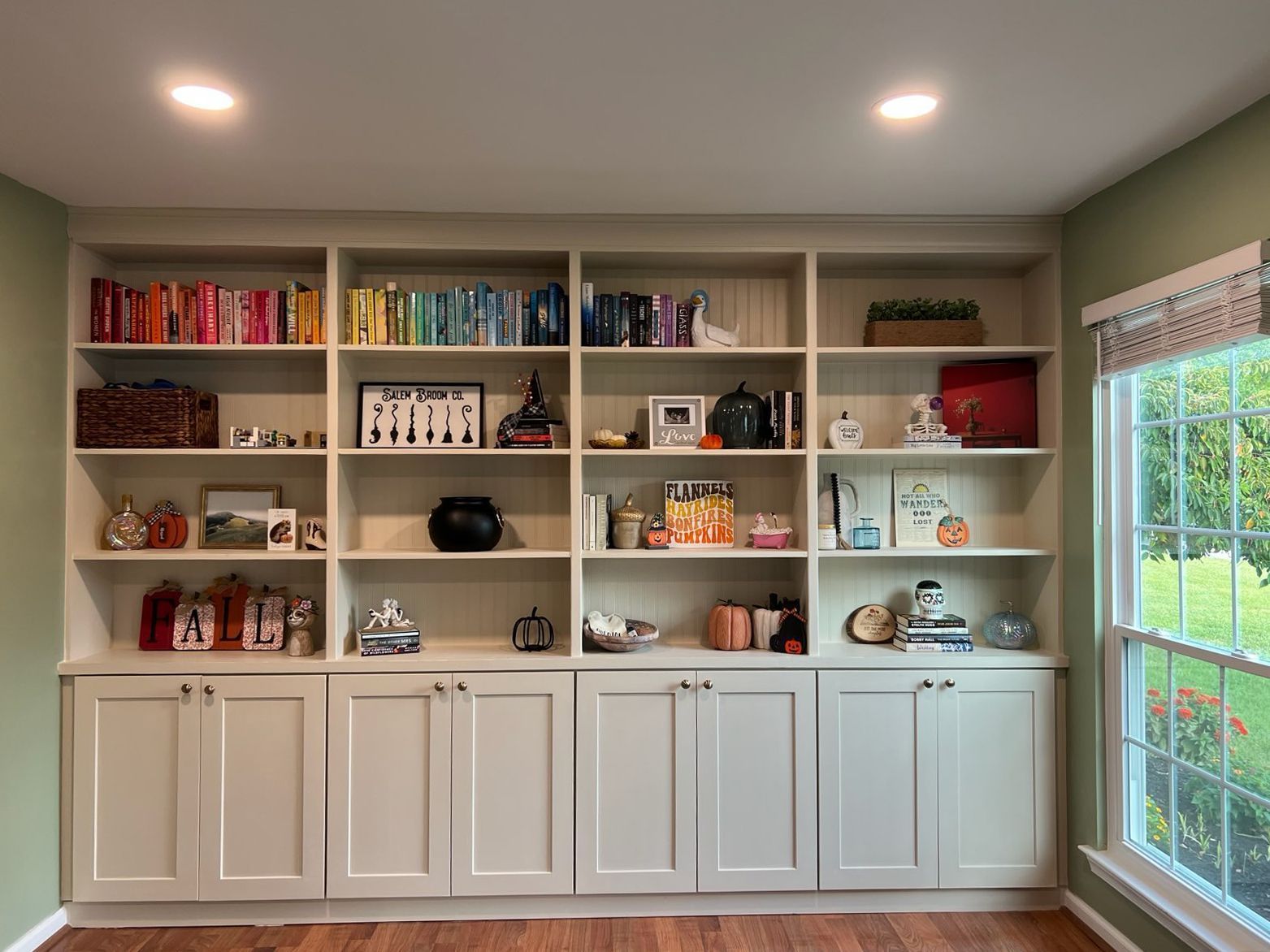Built-in white bookshelves filled with books and decorative items against a pale green wall, next to a window.