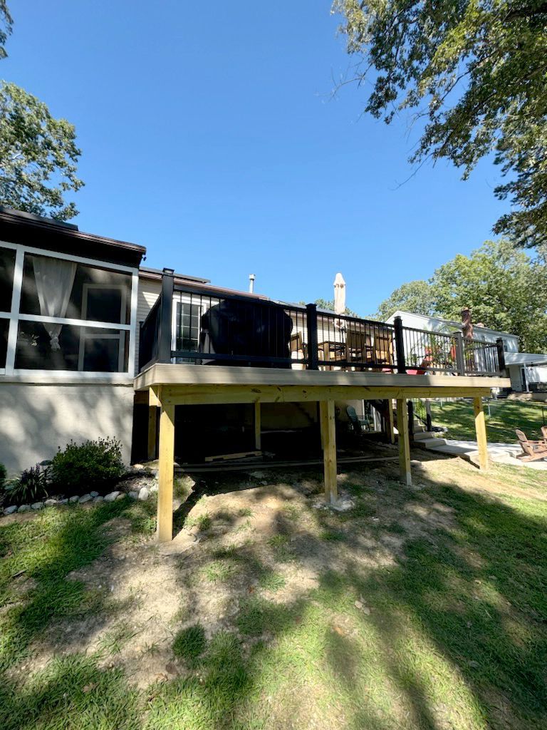 Newly constructed wooden deck with black railing attached to a house; lawn in foreground, trees and sky in the background.