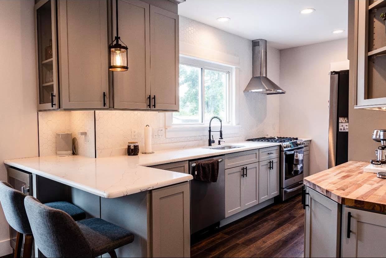 A kitchen with gray cabinets , white counter tops , stainless steel appliances , a sink and a window.