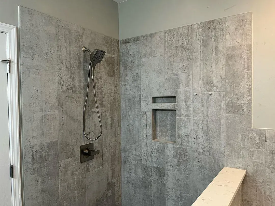 Gray tiled shower with showerhead, built-in shelf, and a partial view of a door.
