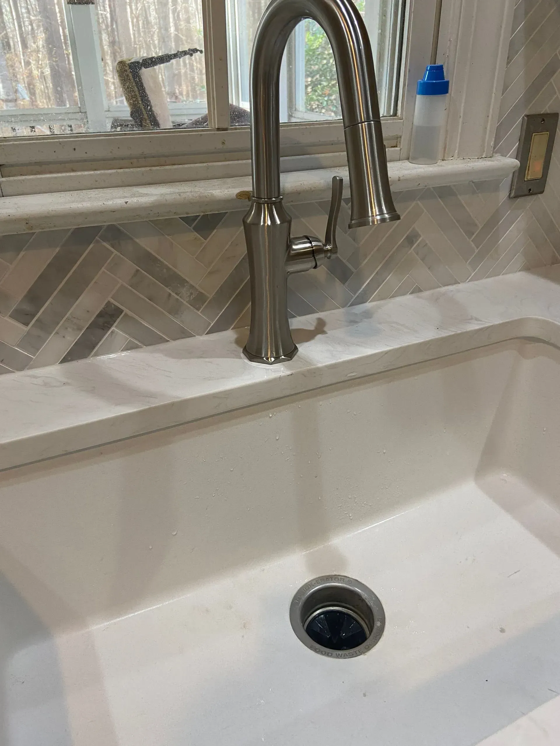 Kitchen sink with stainless steel faucet, white countertop, and gray and beige herringbone tile backsplash.