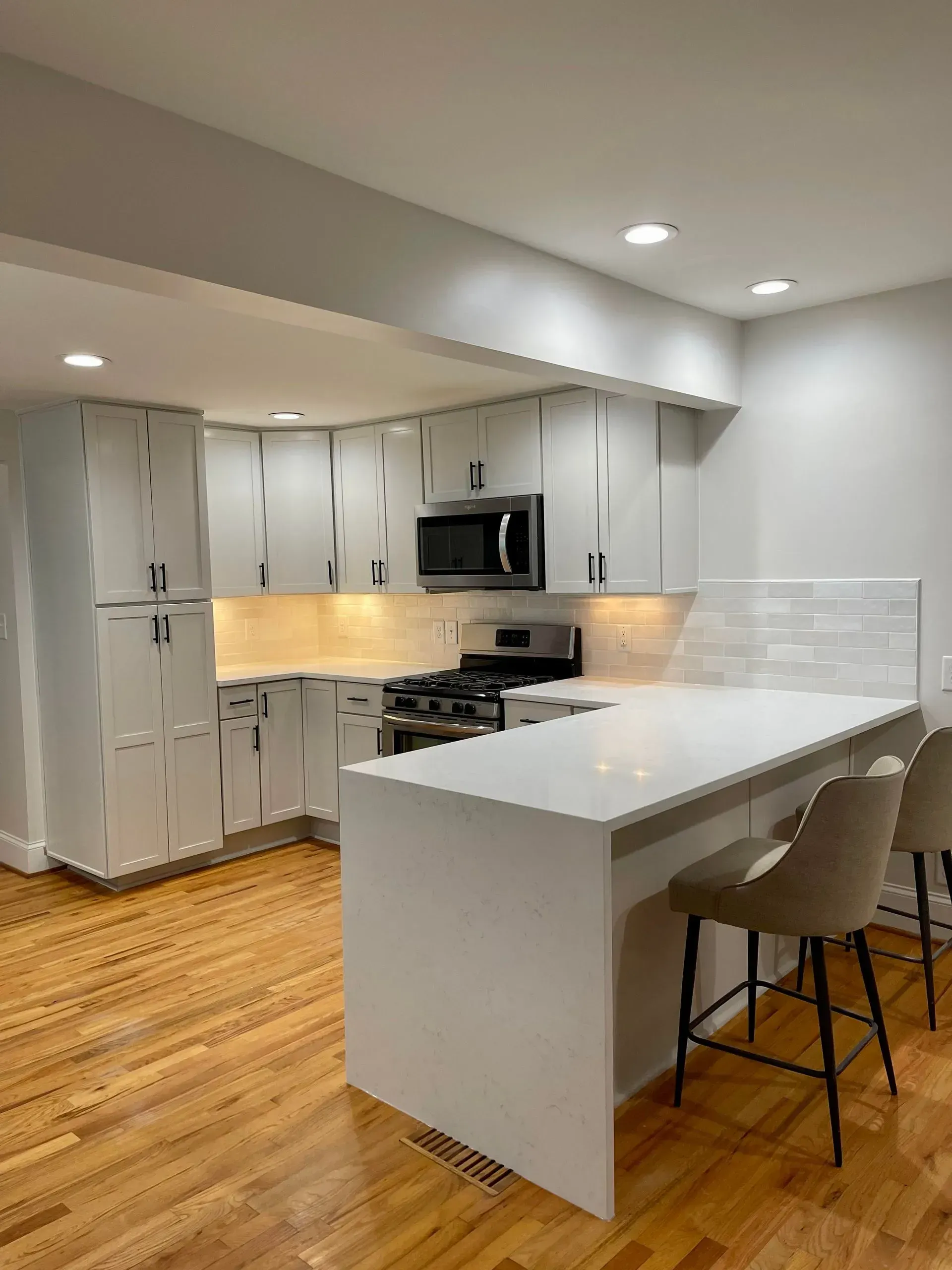 Modern kitchen with white cabinets, countertops, and a breakfast bar. Blonde wood floors, stainless steel appliances.