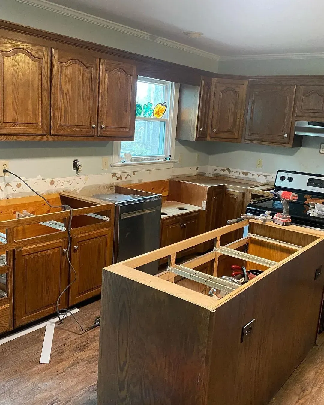 Kitchen undergoing renovation; upper and lower brown cabinets, exposed walls, partially disassembled island, and appliances.