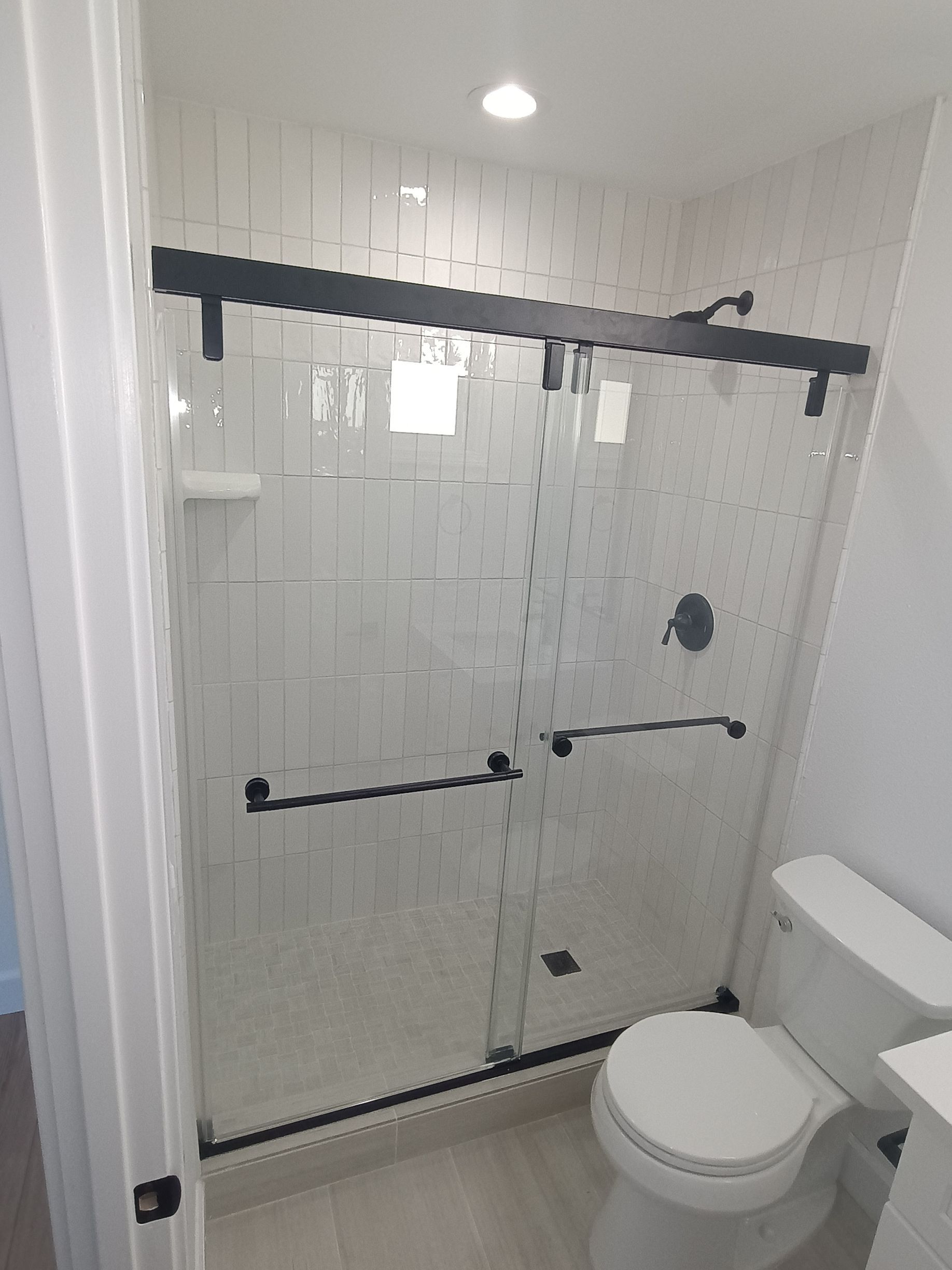 Bathroom with white tile shower, sliding glass doors with black hardware, and a toilet.