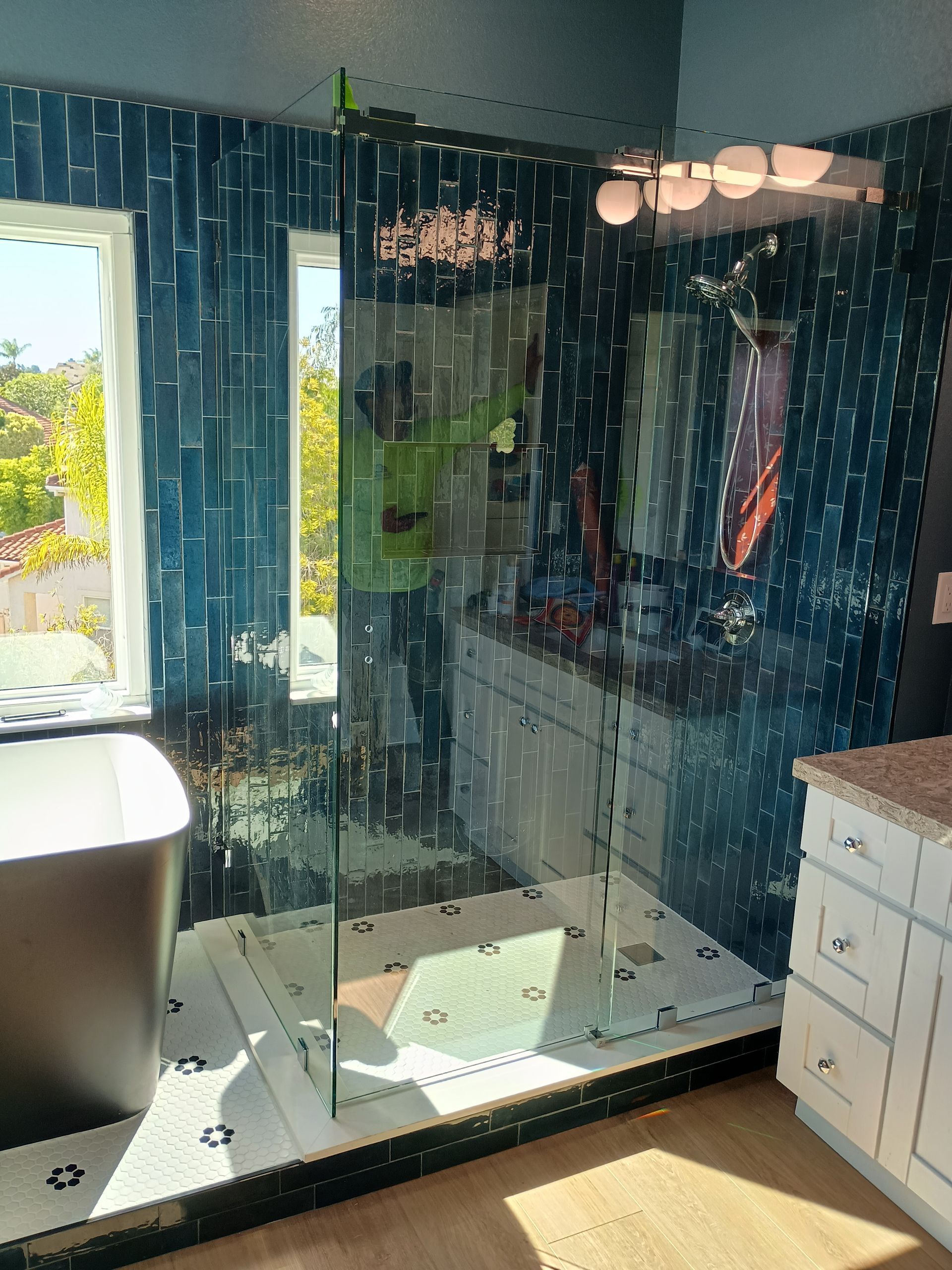 Bathroom with blue tile shower, glass door, white vanity, and tub, sunny window.