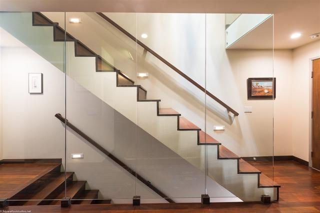 Modern staircase with glass panels and wooden steps; brown handrails, warm lighting, and wood flooring.