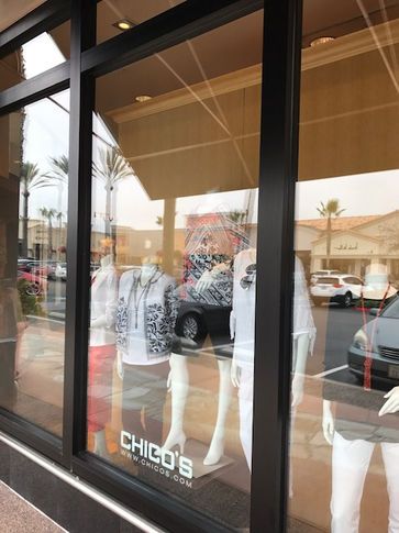 Window display at Chico's clothing store with mannequins wearing outfits. Exterior view with other stores and cars visible.