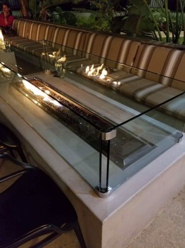 Outdoor seating with a glass-topped table and built-in fireplace. Neutral colors, plants in the background.