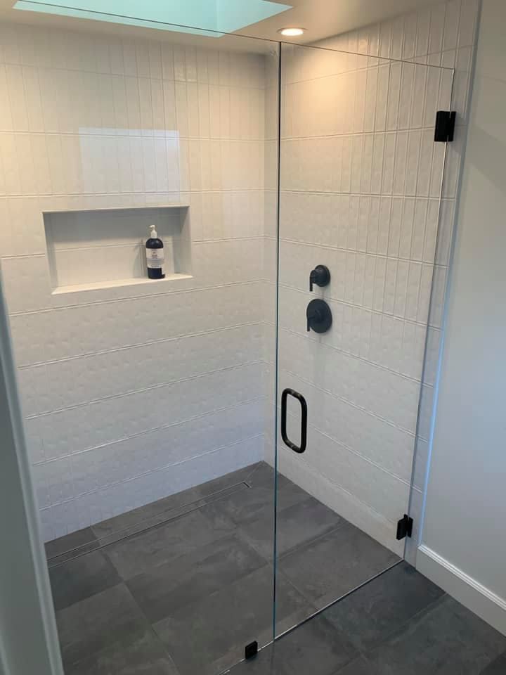 Modern shower with glass door, white tiled walls, gray floor. Black fixtures, built-in shelf.