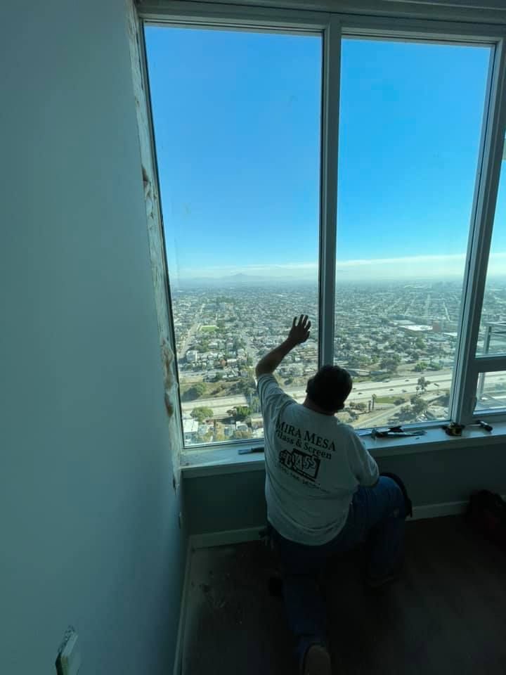 Person kneels near a large window, gesturing toward a cityscape. Clear blue sky.
