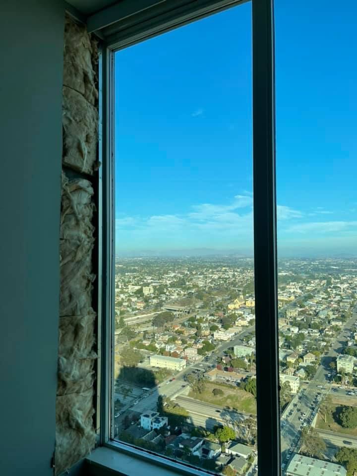 View of a cityscape through a tall window frame. Insulation is visible to the left. Bright blue sky.