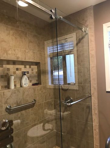 Shower with glass door, grab bars, built-in shelf, tiled walls, and small window with blinds.