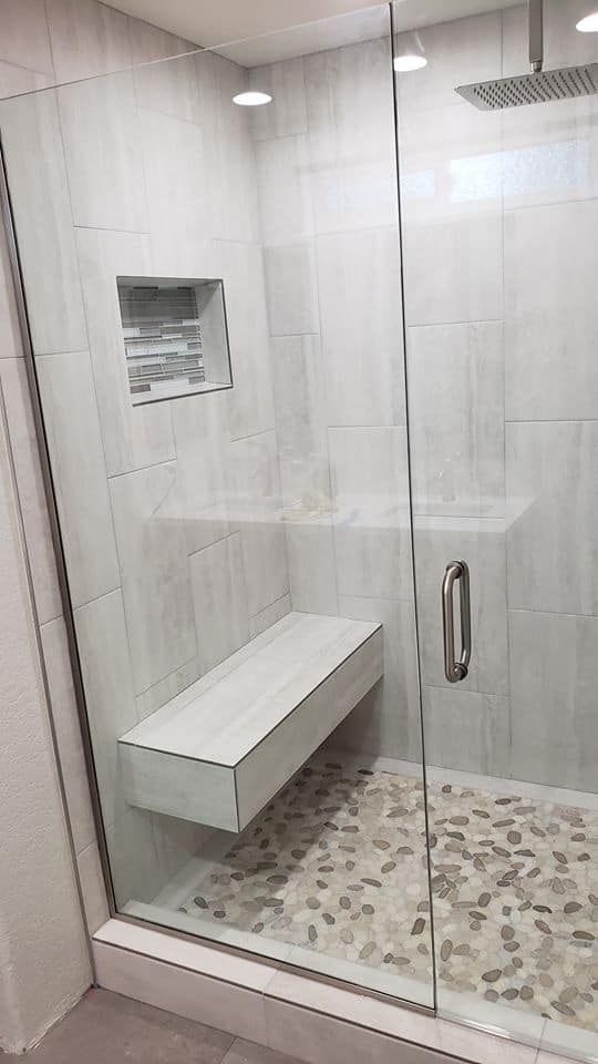 Modern shower stall with glass door, pebble floor, built-in bench, and light gray tiled walls.