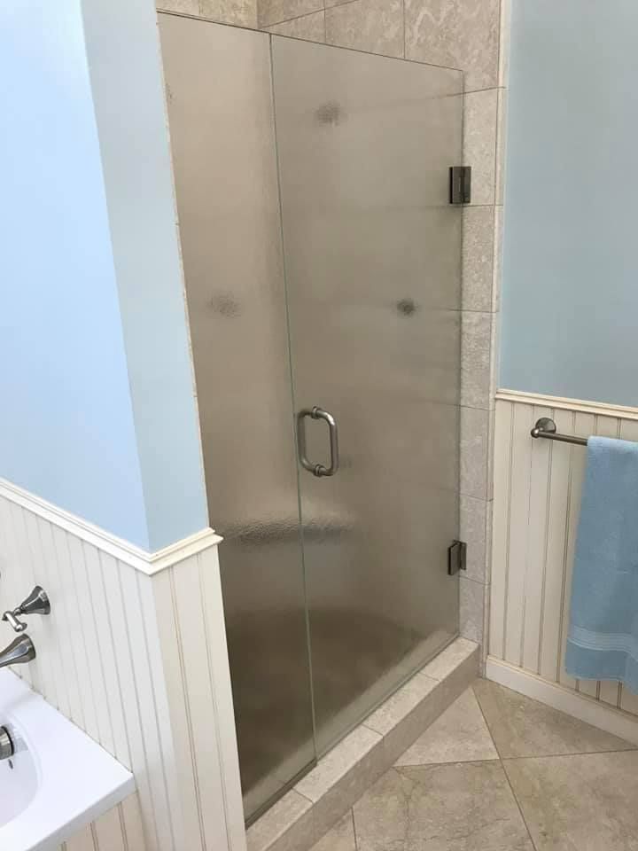 Shower with glass door, brushed nickel handle and hinges, set in bathroom with pale blue walls and white wainscoting.