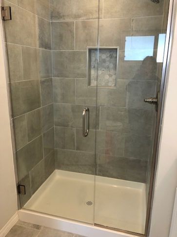 Gray-tiled shower with glass door and white base. Includes a recessed shelf and silver hardware.