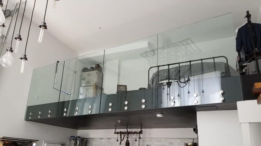 Loft bedroom with glass railings over a dark gray structure, white walls, and industrial-style lighting.