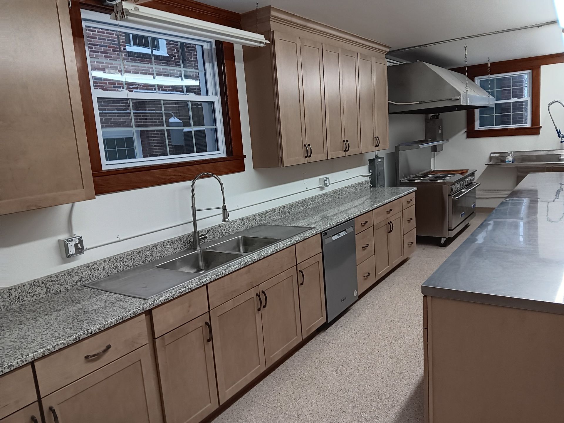 Long commercial kitchen with stainless counters, sinks, wood cabinets, and a large window