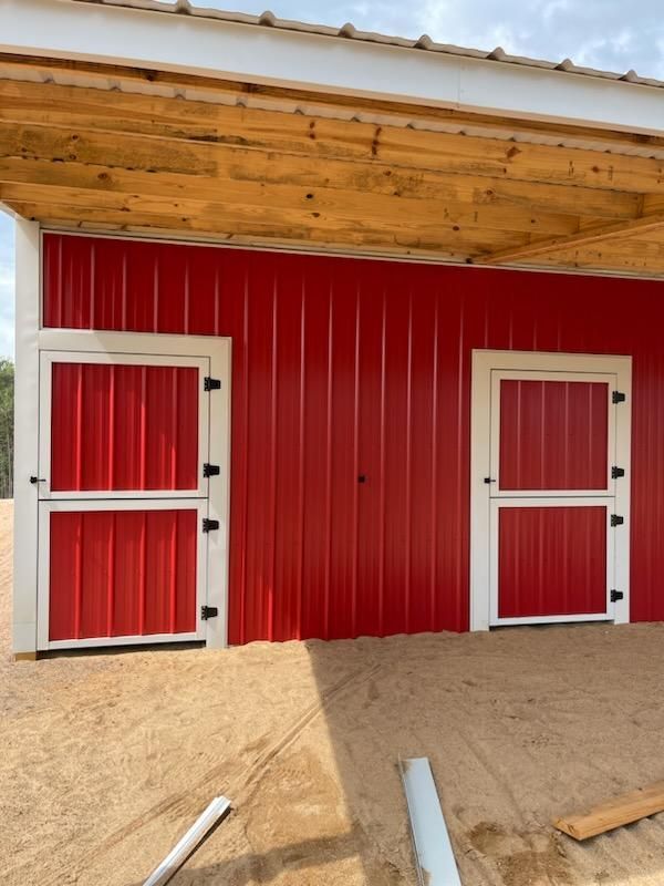 Two red Dutch doors with white trim and black hinges on a red metal barn under a wooden overhang.