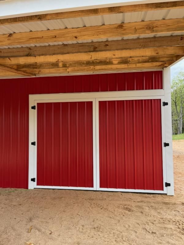 Double red metal barn doors with white trim and black hinges beneath a wooden overhang.