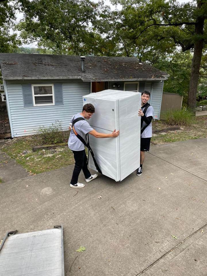 Two men are carrying a refrigerator in front of a house.