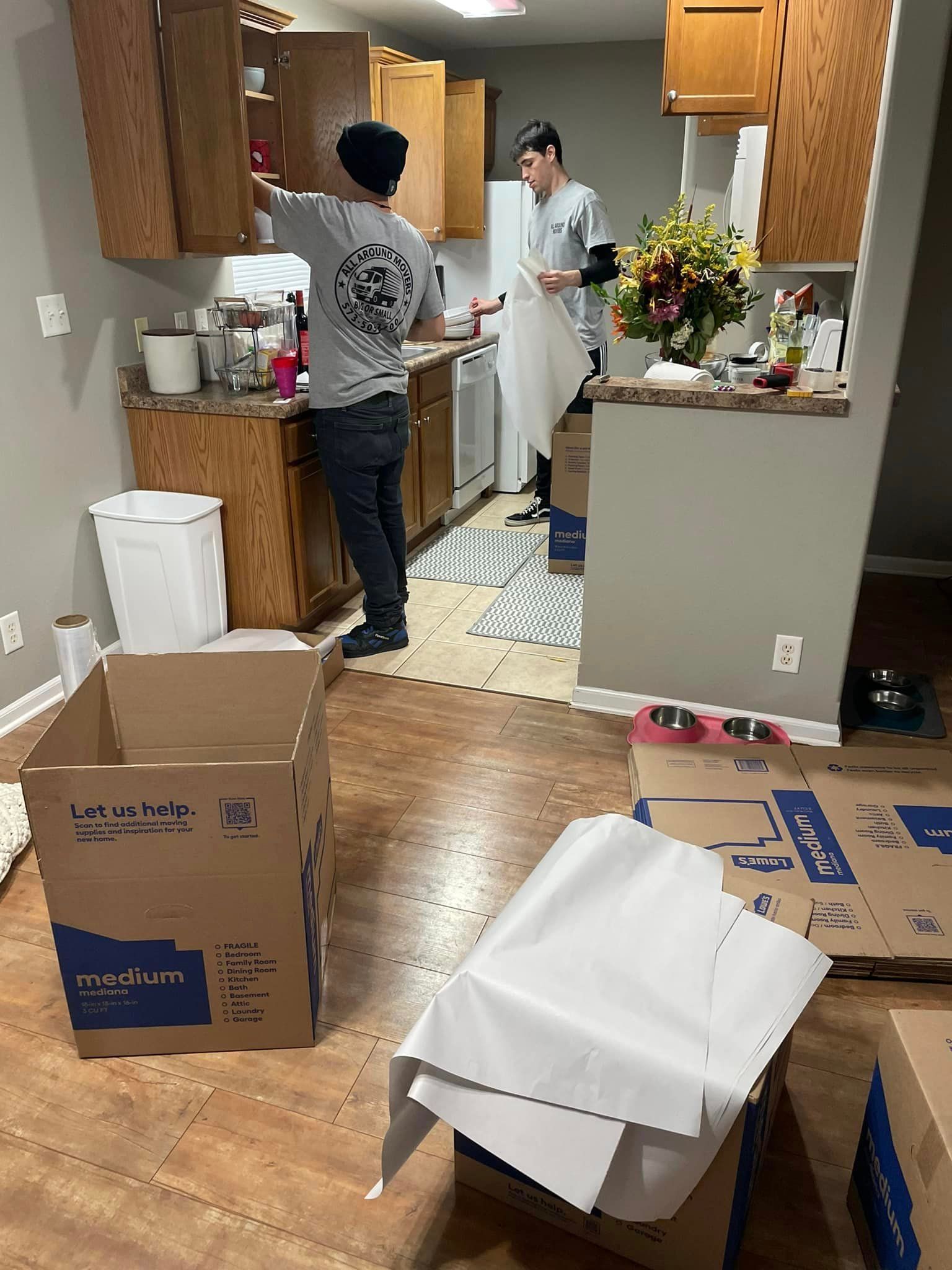 Two men are standing in a kitchen surrounded by cardboard boxes.