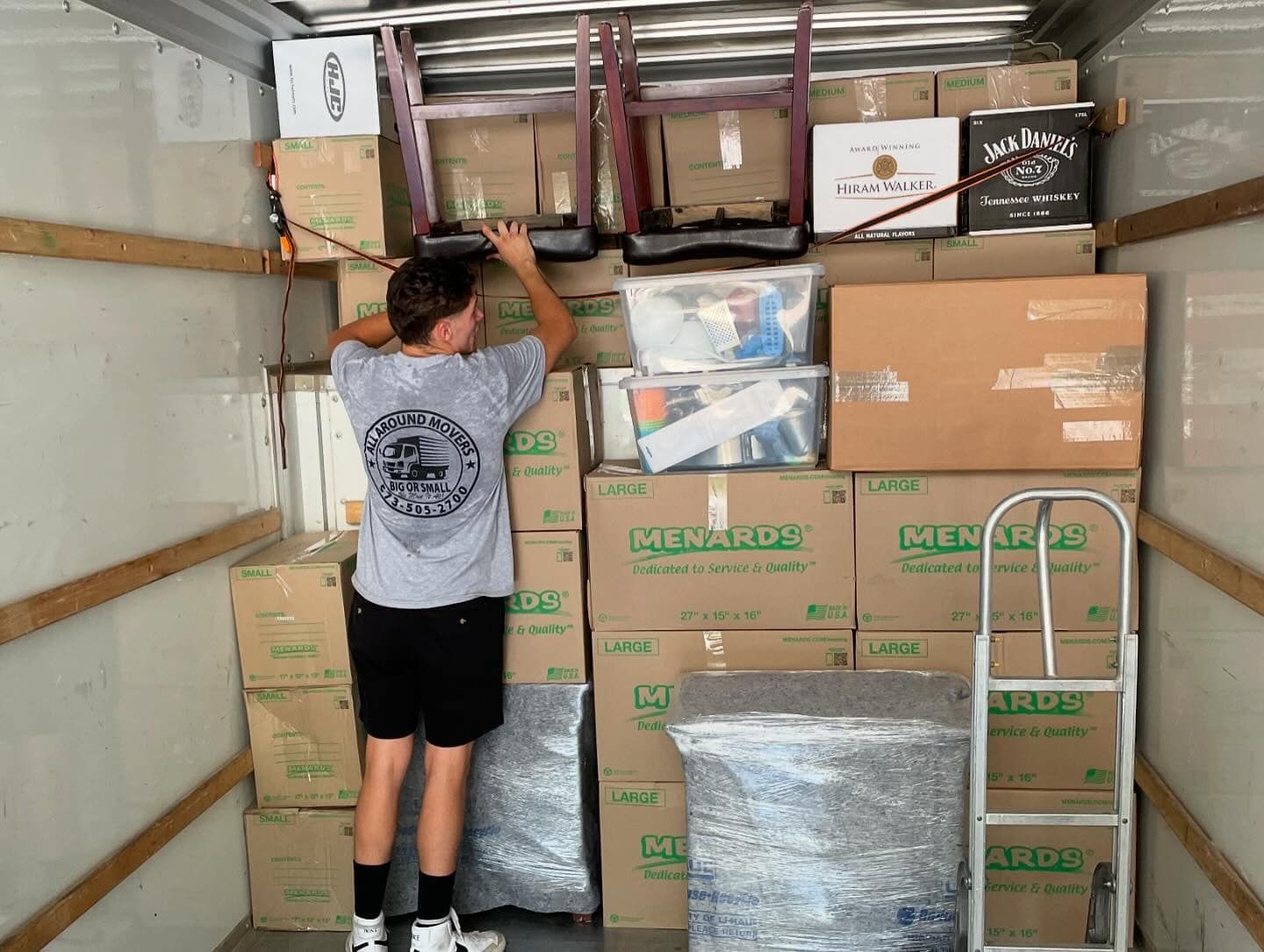A man is standing in a truck filled with boxes and chairs.