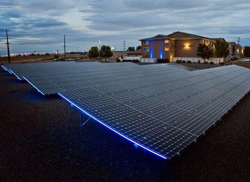 Large solar panel array in a parking lot, illuminated by blue lights, with a hotel in the background.