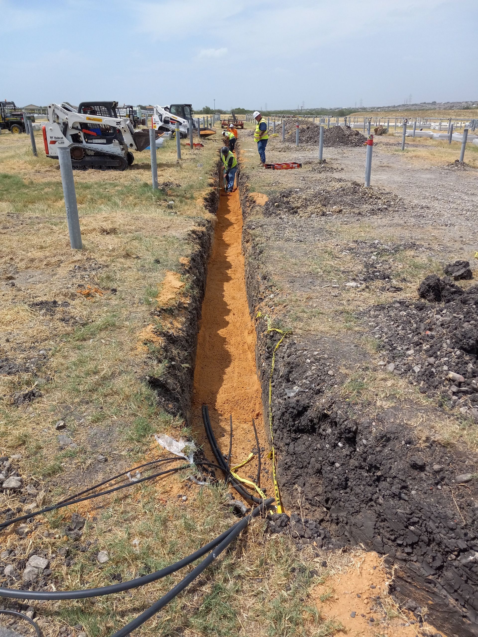 Construction workers in a trench, laying cables. Outdoors with a field, sunny, and poles for solar panels.