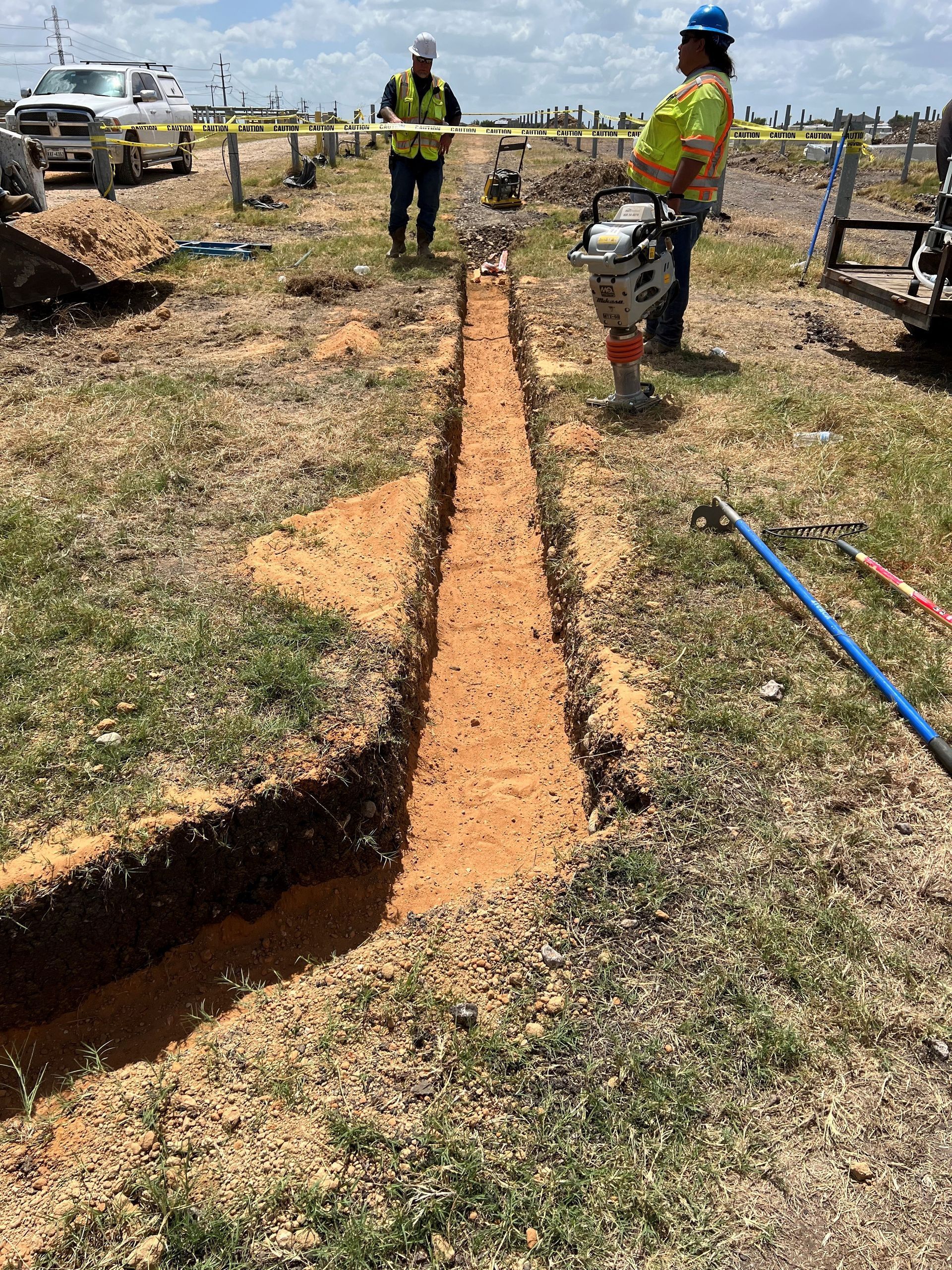 Two workers compact soil in a trench with equipment outdoors.