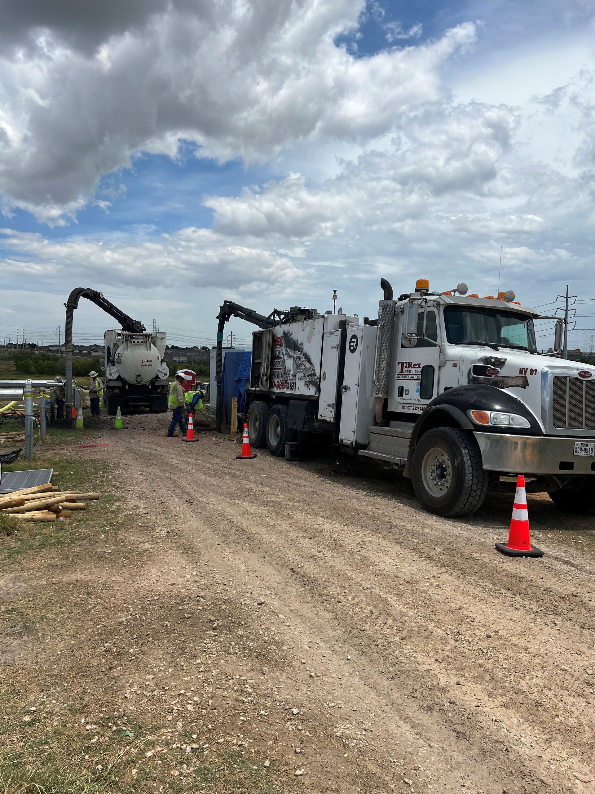 Two industrial trucks extracting material on a gravel road, workers in safety vests nearby under a cloudy sky.