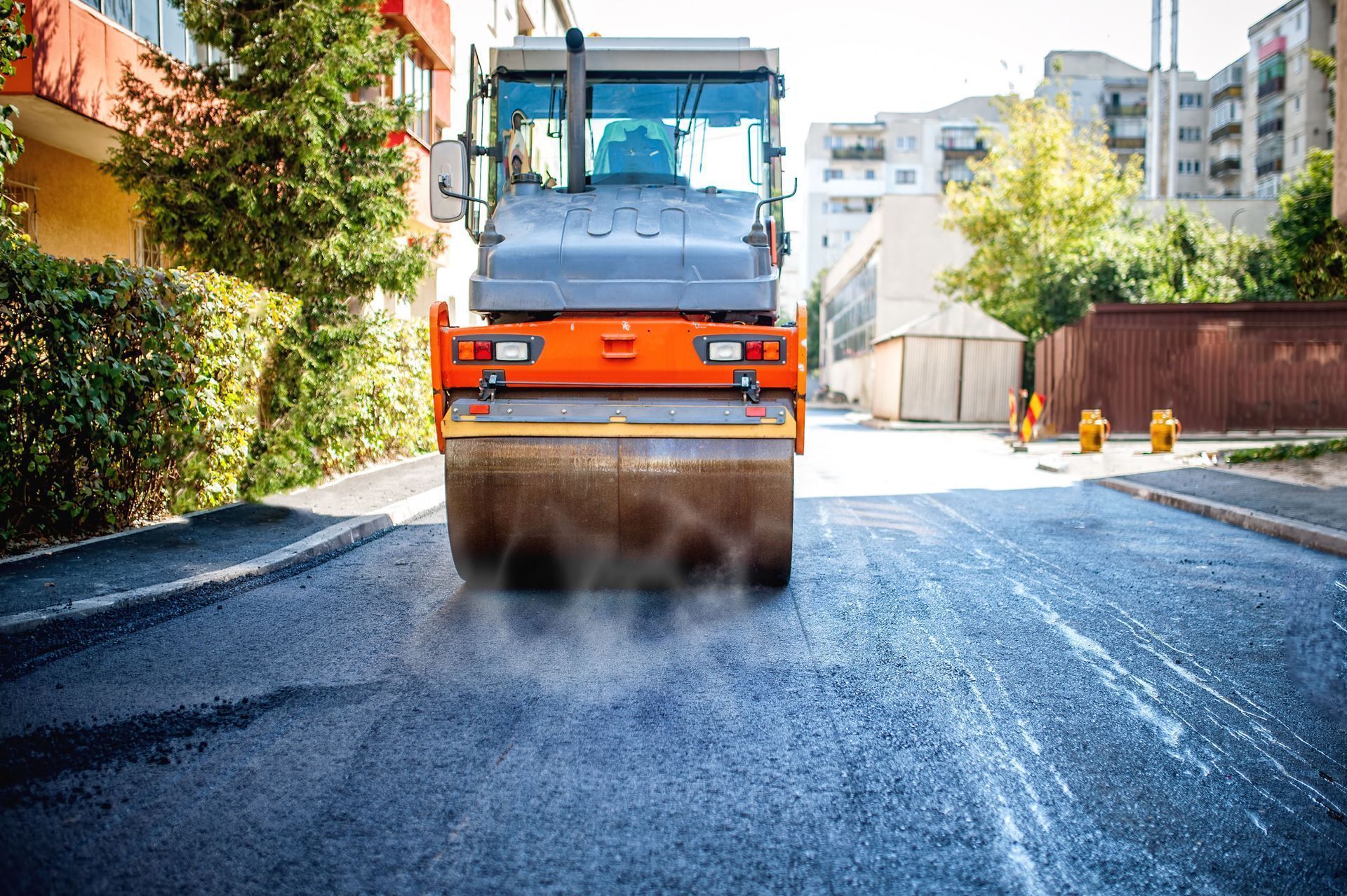 Road roller compacting fresh asphalt on a city street, orange and gray machinery.