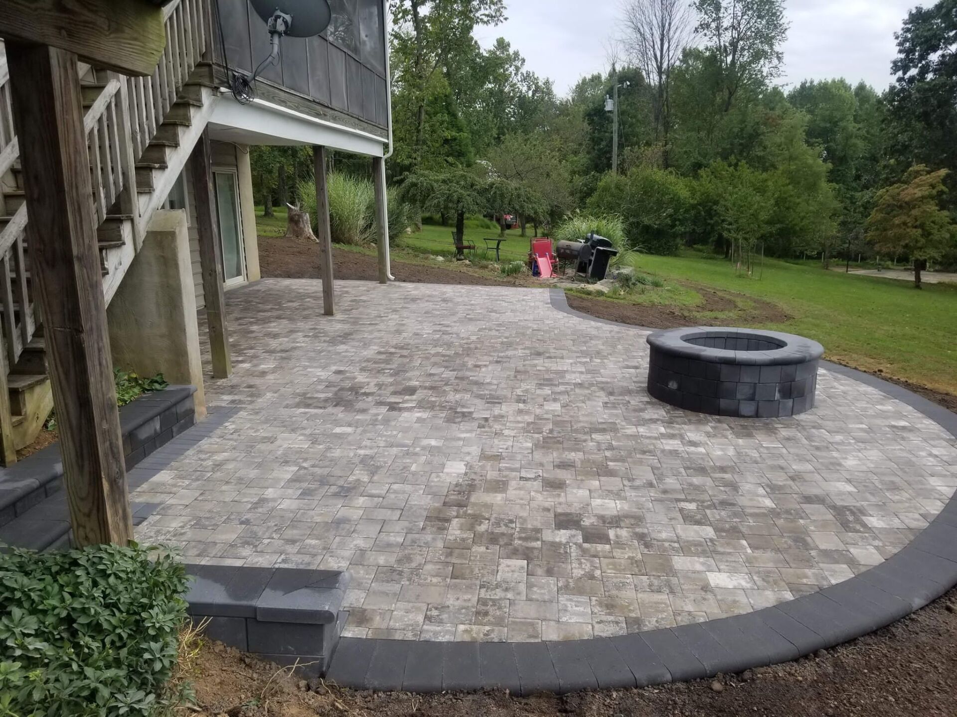Brick patio with fire pit, steps, and house under deck, surrounded by grass and trees.