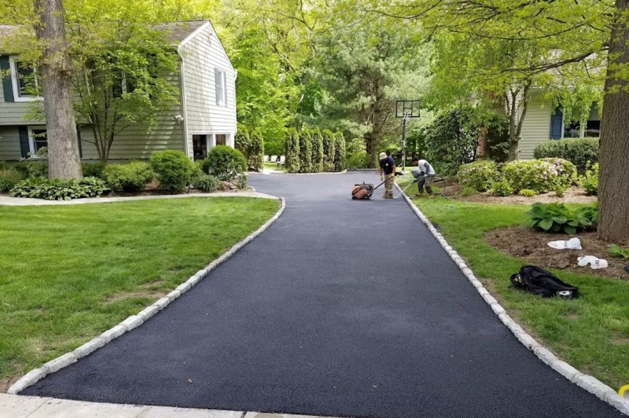 Newly paved asphalt driveway with workers; green lawns and trees surround a house.