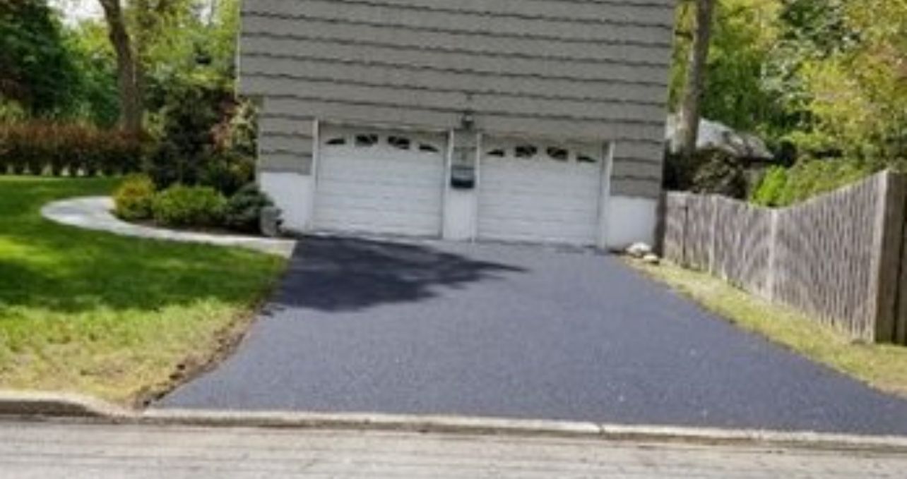 Asphalt driveway leading to a two-car garage with white doors, flanked by grass and a wooden fence.