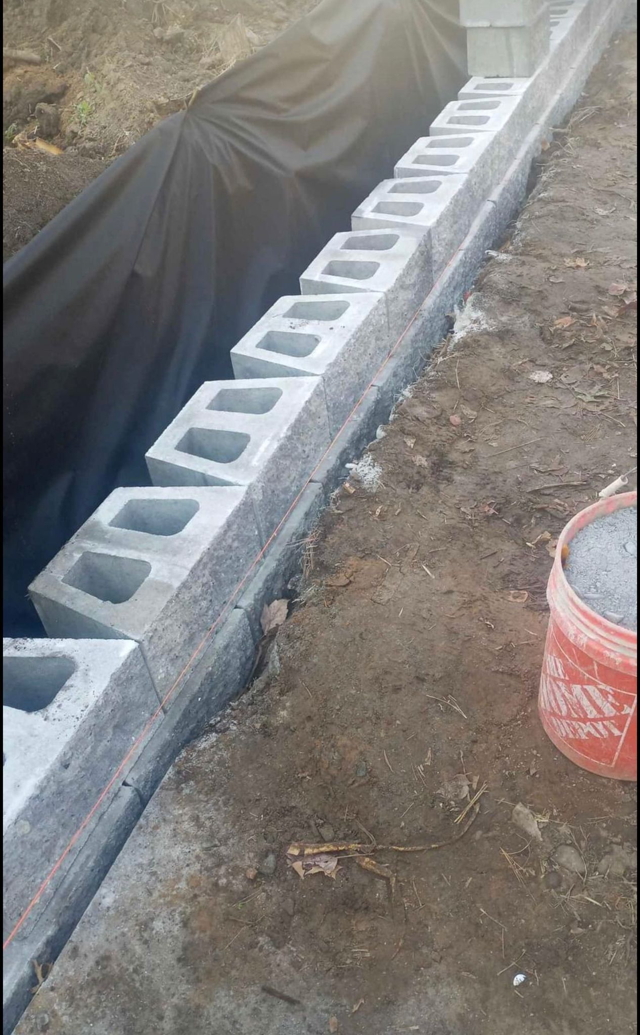 Concrete blocks forming a wall in a trench, next to soil and a black liner. A red bucket sits nearby.