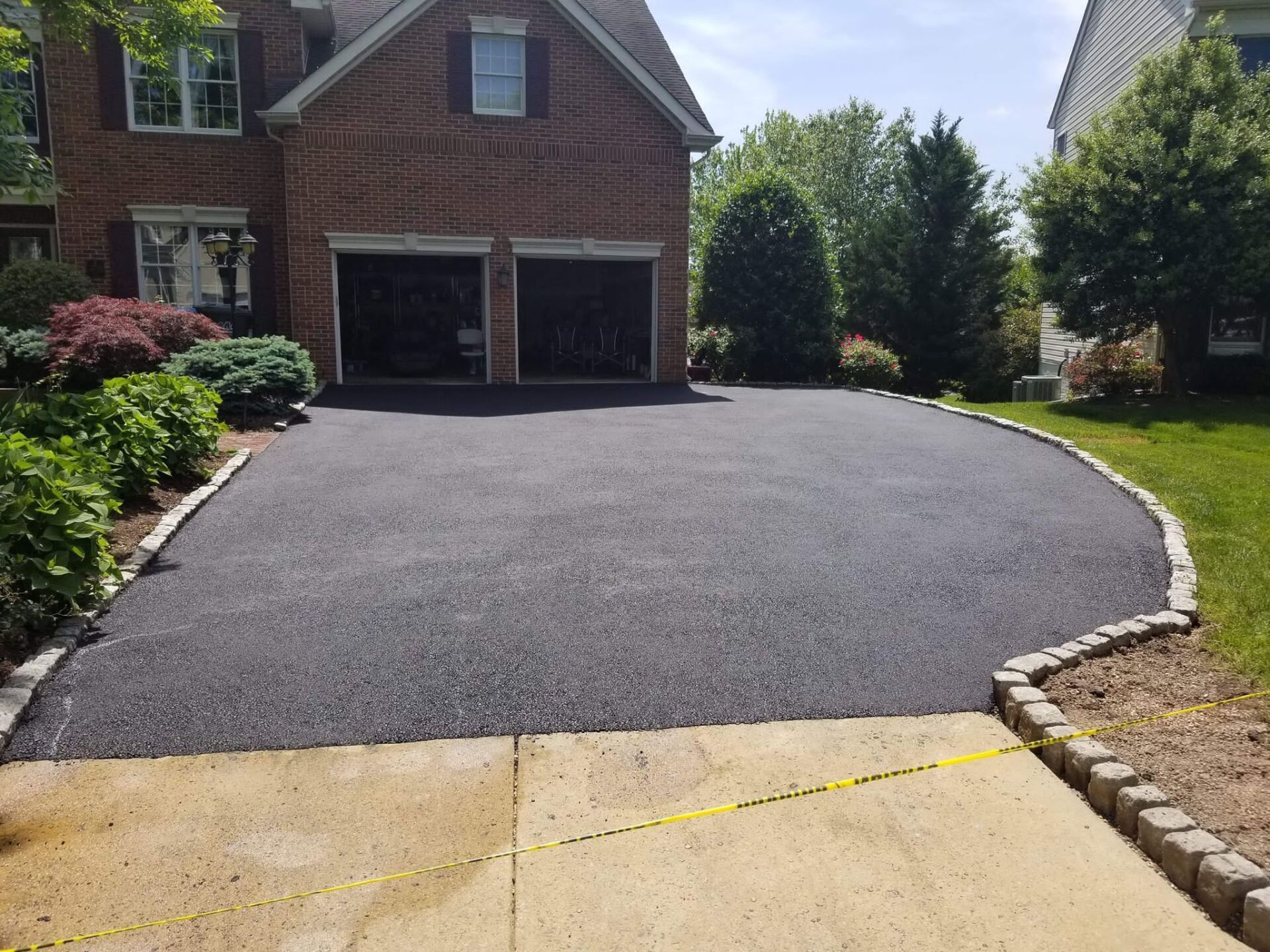 Driveway freshly paved in black asphalt, in front of a brick house with garage and landscaping border.
