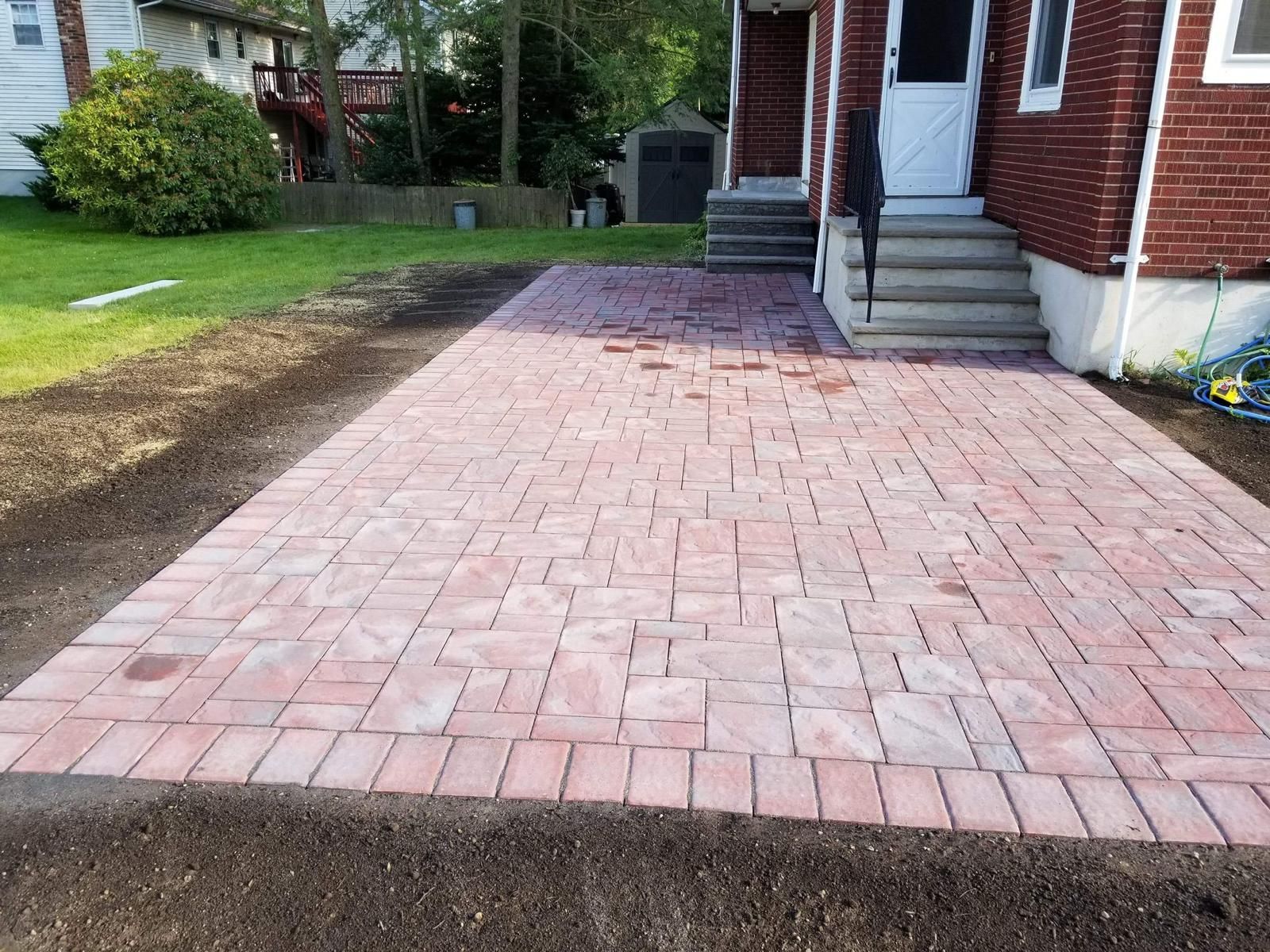 Red brick patio next to a red brick house with steps leading to a white door, on a grassy lawn.