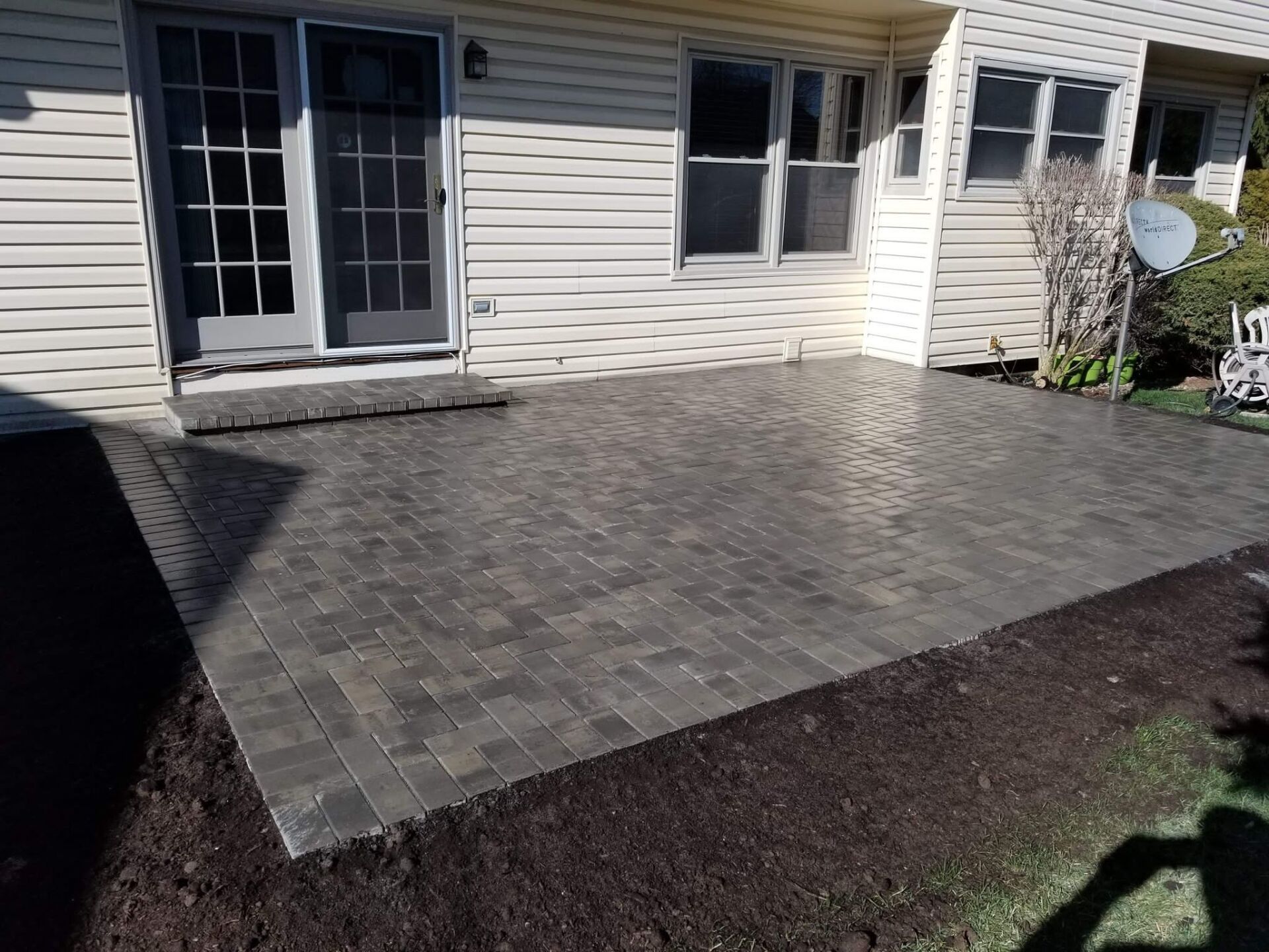 Brick patio extends from a house with a door and windows, surrounded by dirt and grass.