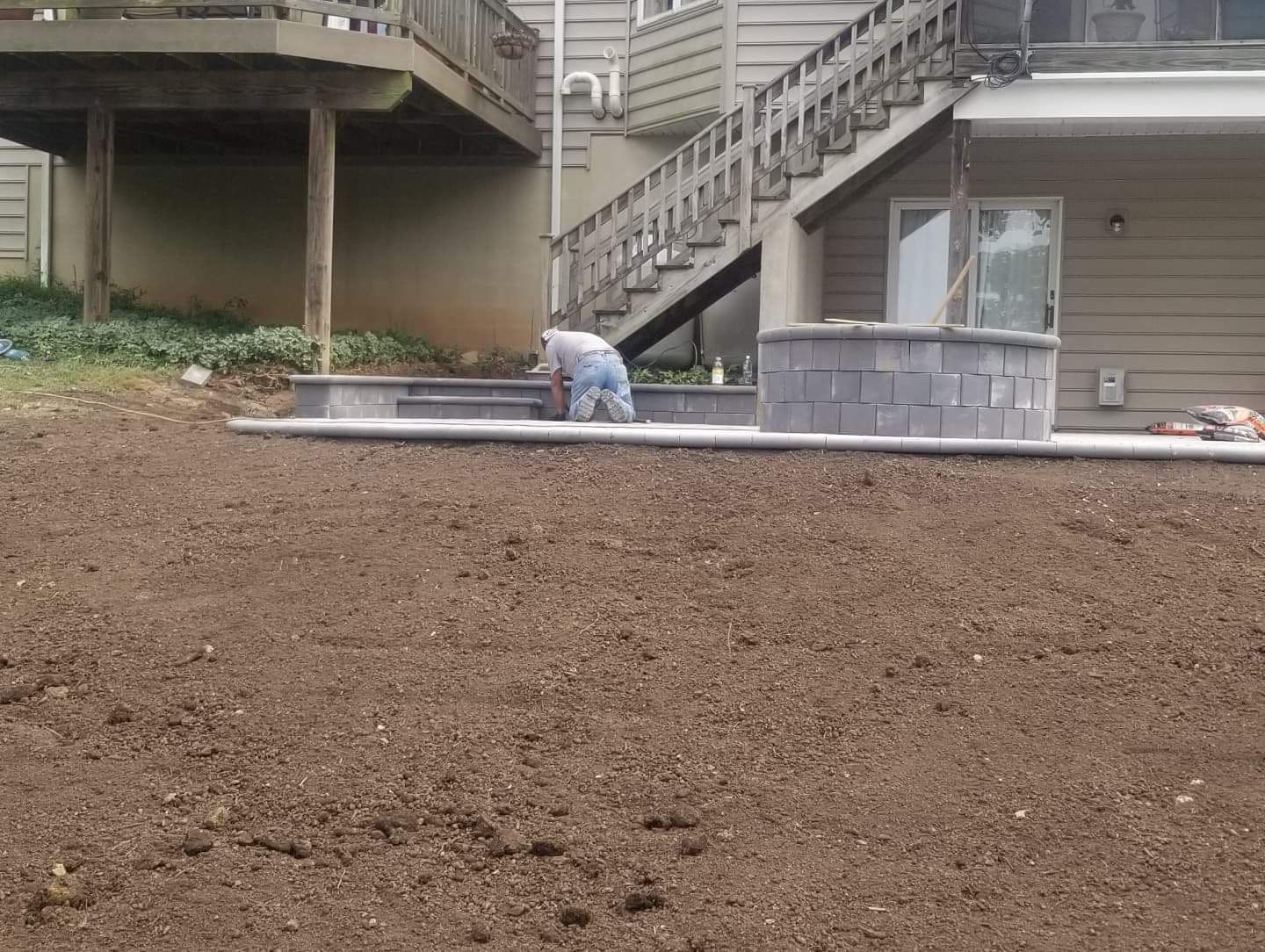 Man working on a newly built patio with a fire pit, next to a house with stairs and a deck.