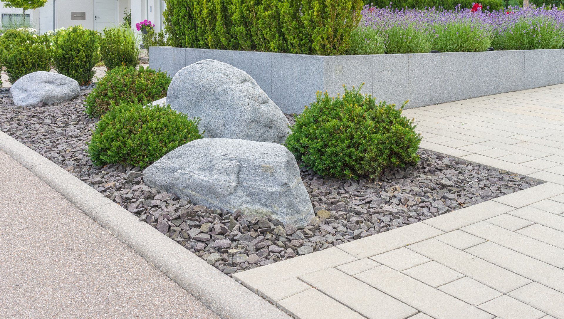 Landscaped garden bed with large rocks, green bushes, and dark mulch, adjacent to a paved walkway.