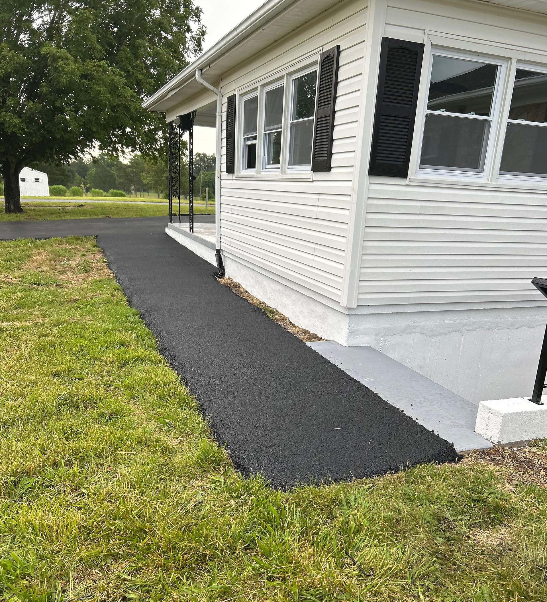 Asphalt pathway next to a white house with black shutters, on a grassy lawn.