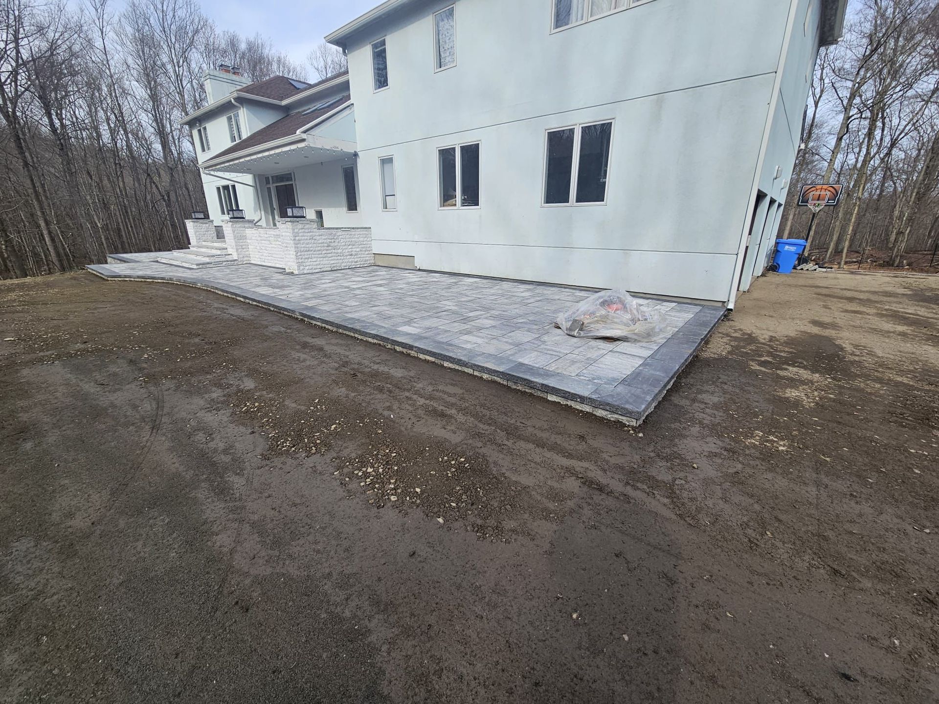 A two-story house with a newly constructed stone patio. Dirt and gravel in the foreground.