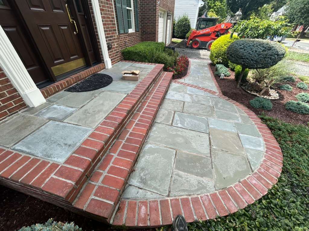 Stone walkway and steps with brick borders lead to a front door with landscaping, including a small excavator in the background.