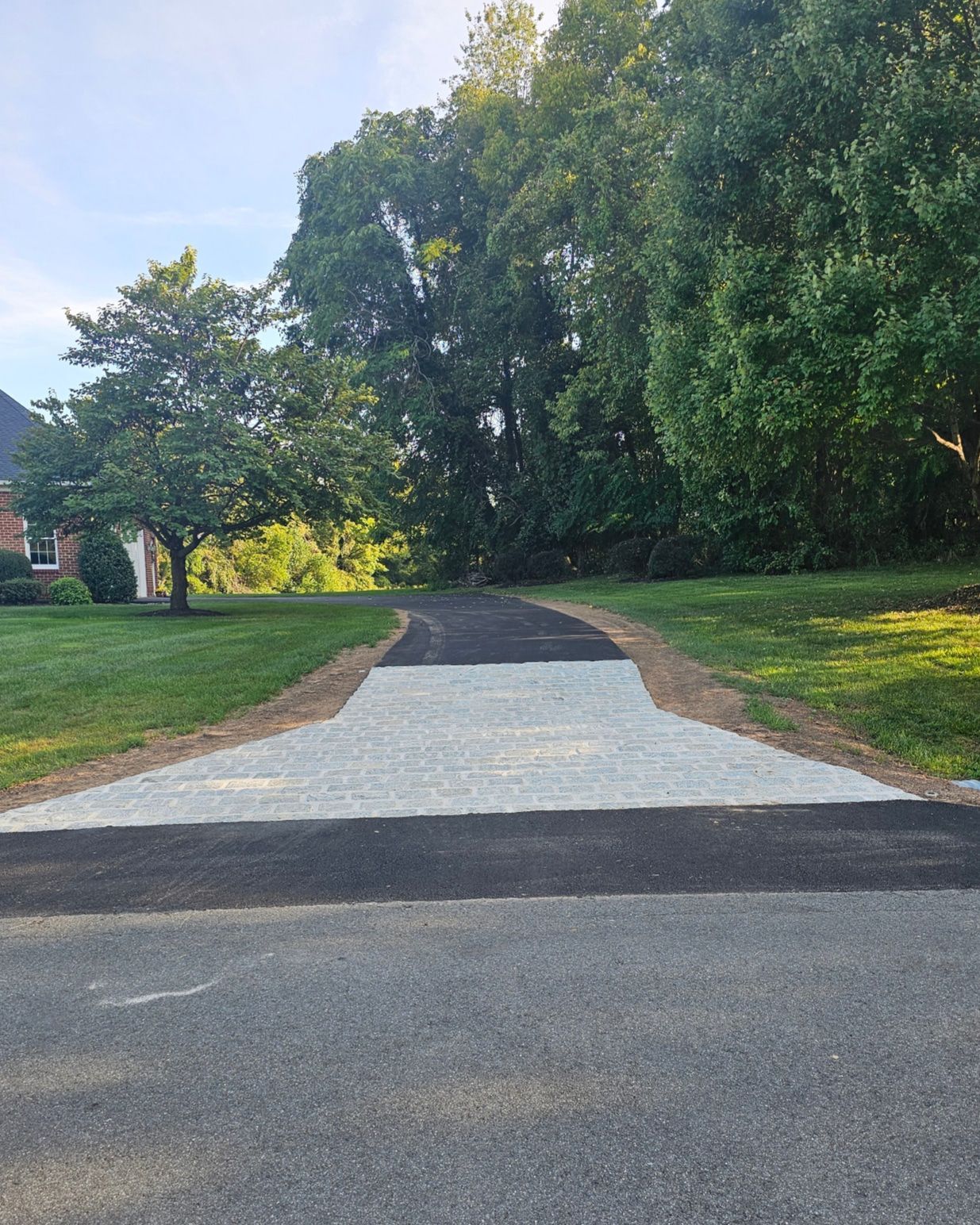 Asphalt driveway with gravel extension, leading into trees, on a sunny day.